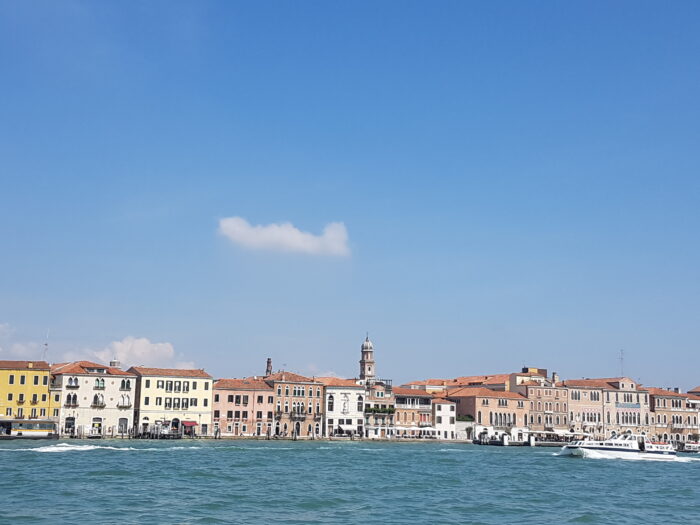 Happy Life: Colorful historic buildings along the Venice waterfront with a church tower in the background and boats on the water.