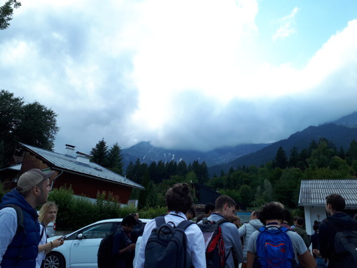 Group of hikers with backpacks gathered in front of alpine cottages with misty mountains and dense forests in the background.