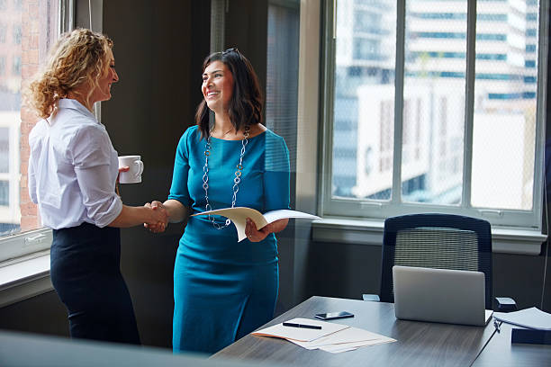 Two businesswomen shaking hands and empowering women with smile after a meeting in a modern office.